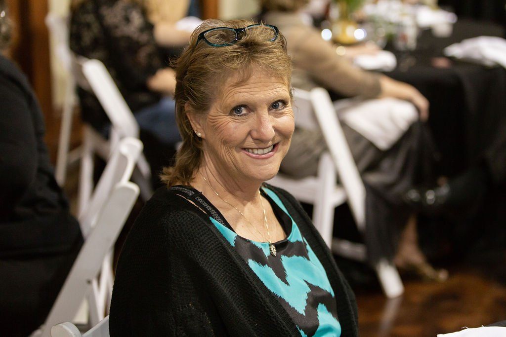 A smiling person with glasses perched on their head, wearing a teal patterned top and black cardigan in a dining hall.