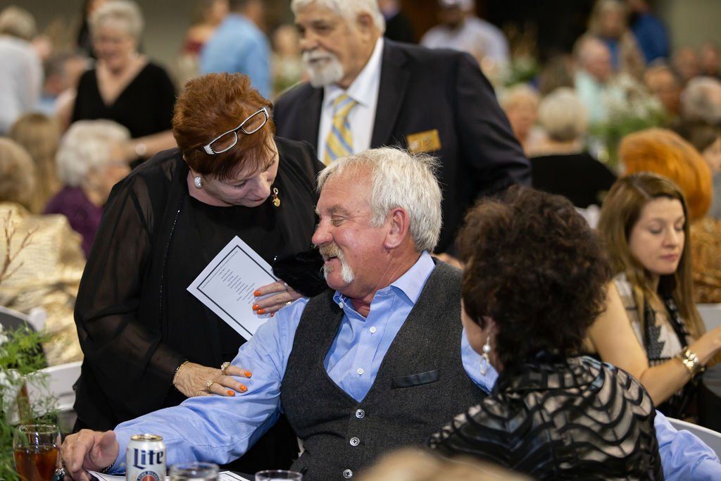 A woman in black leans over to talk to a man in a blue shirt and vest at a crowded, brightly lit banquet hall event.