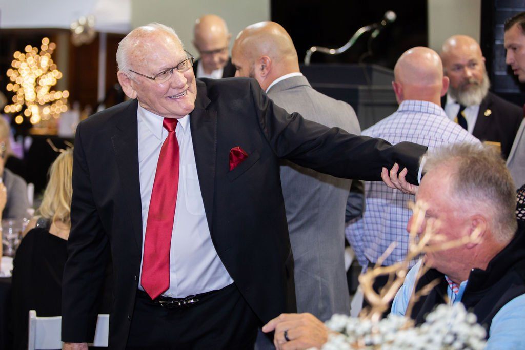 A smiling individual in a suit and red tie gestures toward someone seated at a table during an indoor event.