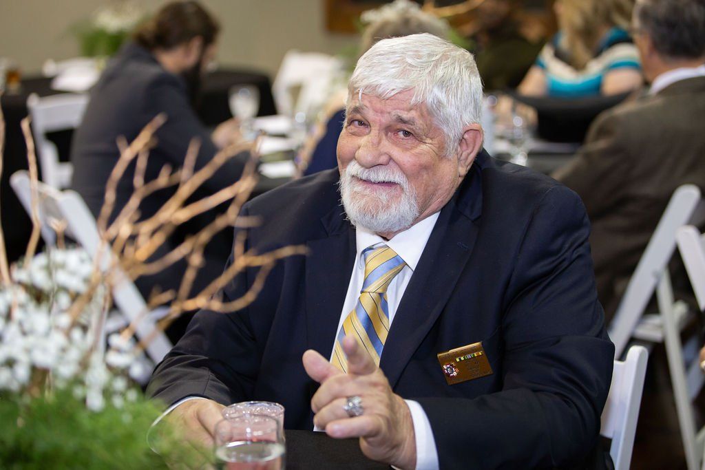 A man in a suit and tie smiling and gesturing with his finger, seated at a table in a bright event space.