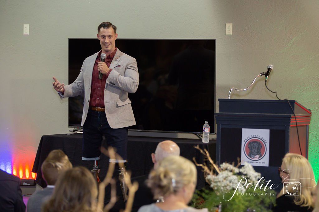 A speaker stands at a podium in a room, holding a microphone and gesturing while addressing an audience.