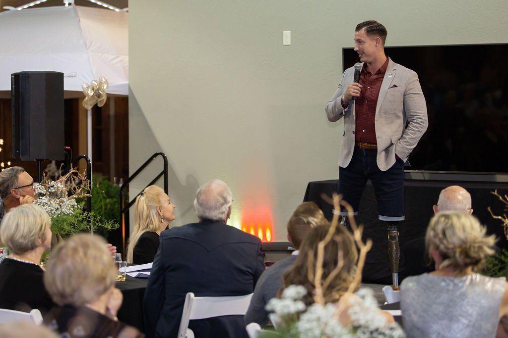 A person with a prosthetic leg stands and speaks into a microphone before an audience in a bright event space.