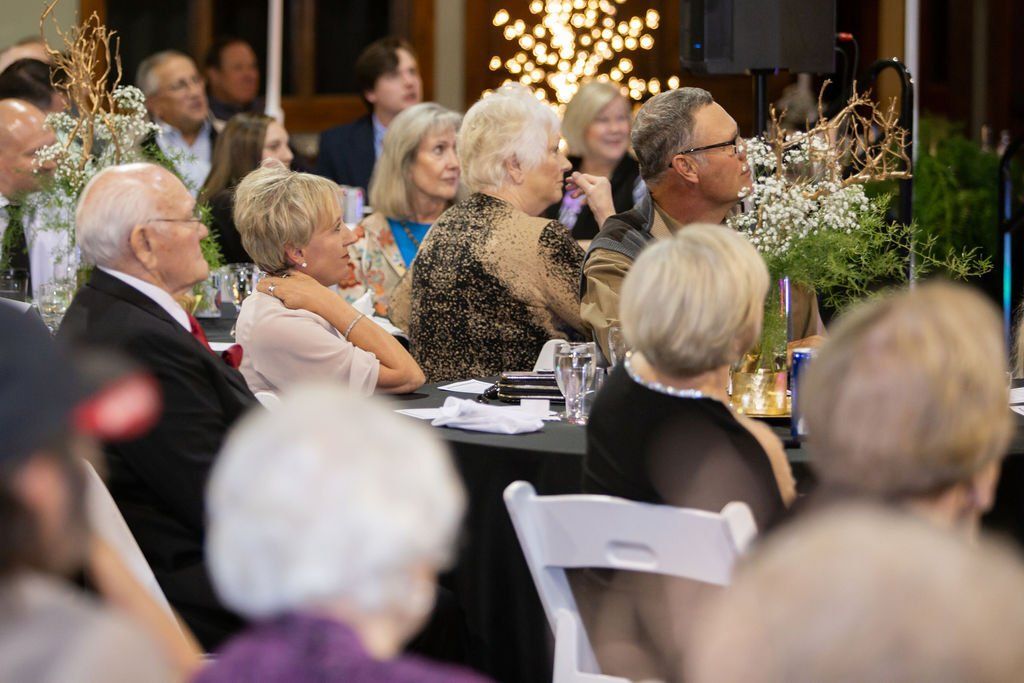 People seated at tables at an event, focusing their attention toward a speaker or performance in the distance.