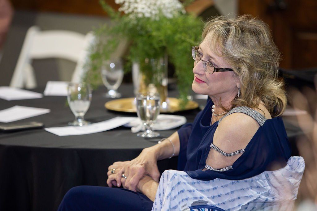 A person in a blue sleeveless top sitting at a table with a black tablecloth, looking to the side with a gentle smile.