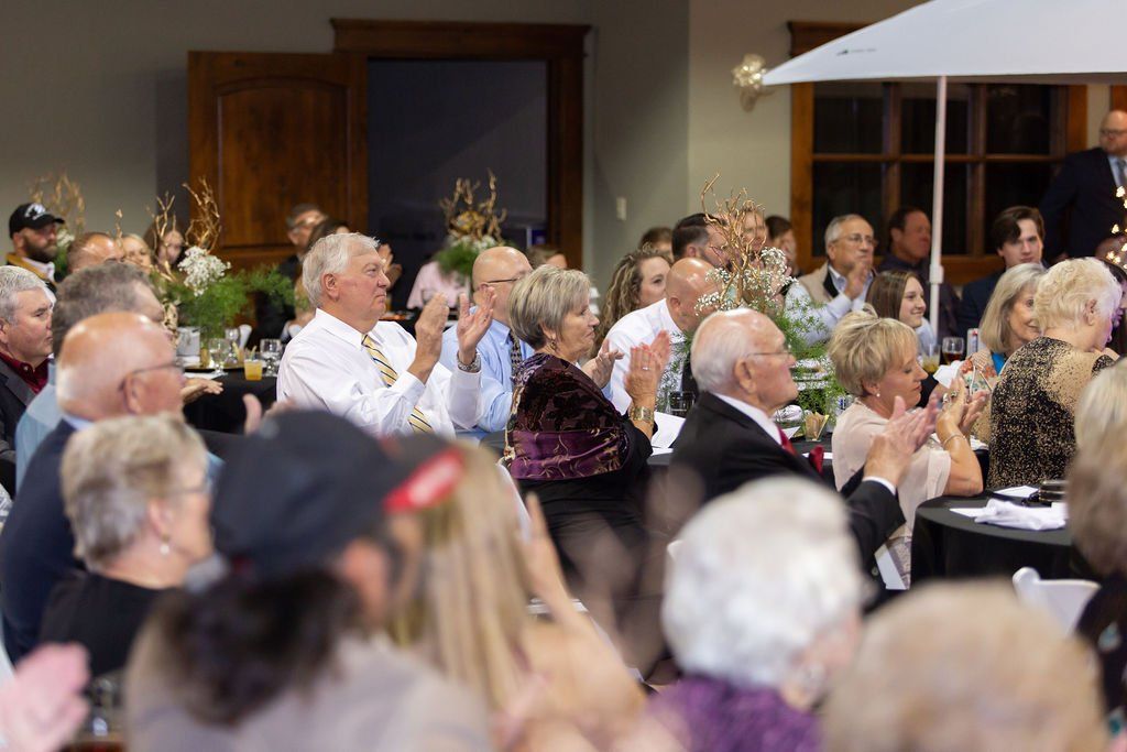 An audience claps during a formal indoor event, with people seated at tables in a dimly lit room.