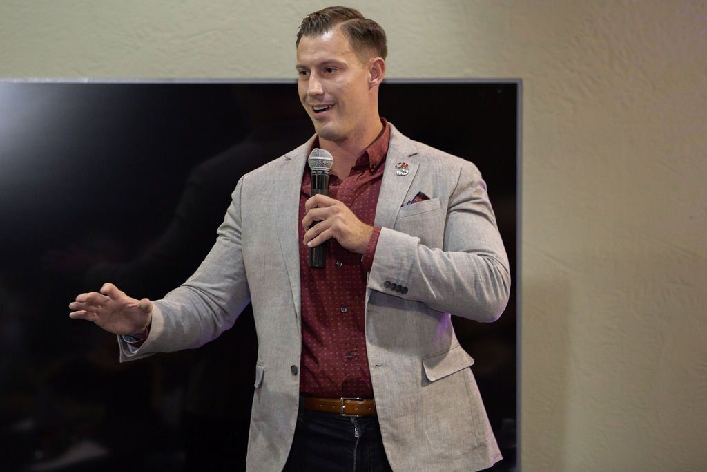 A person in a light blazer and maroon shirt speaks into a microphone while gesturing in front of a dark screen.