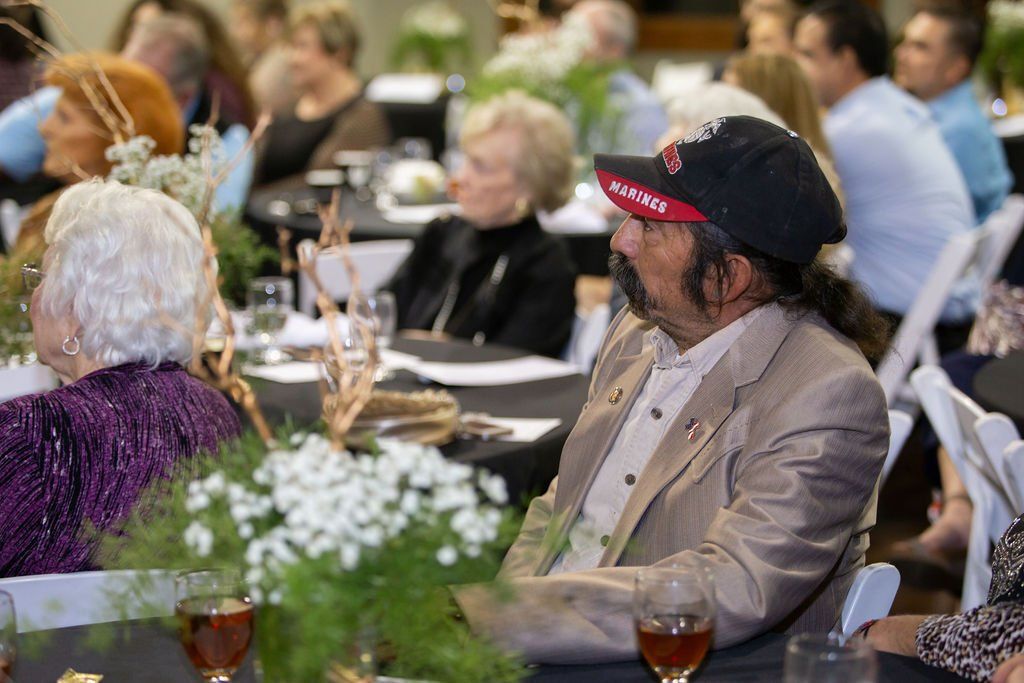 People seated at round tables during an event, featuring a foreground floral arrangement and a man in a tan blazer.
