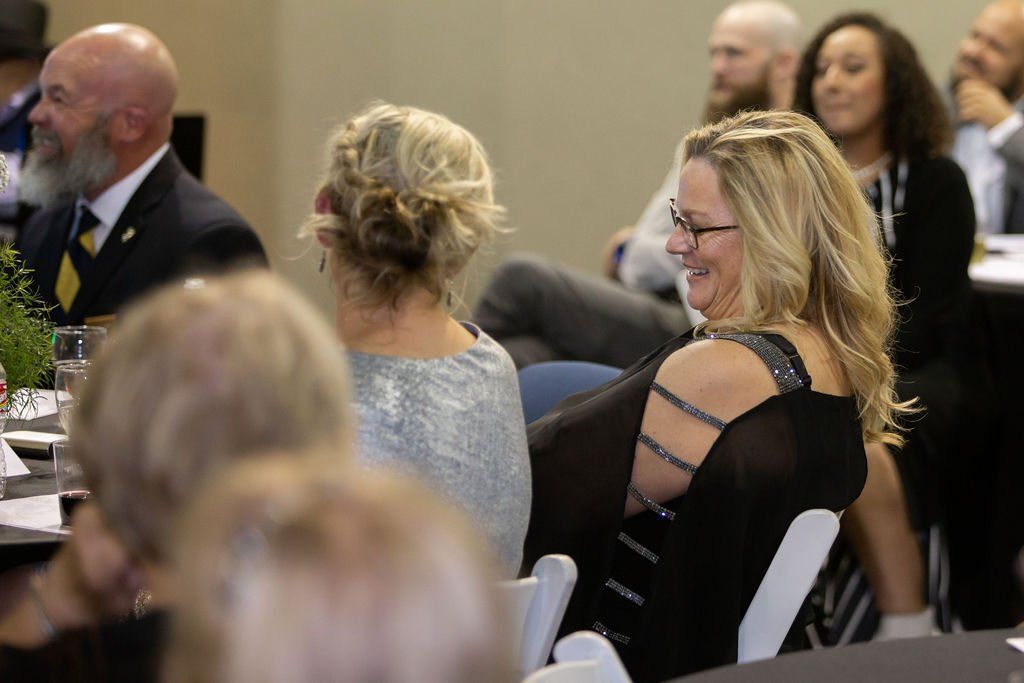 Attendees in formal attire sit at tables in a conference room, listening and smiling during an event.