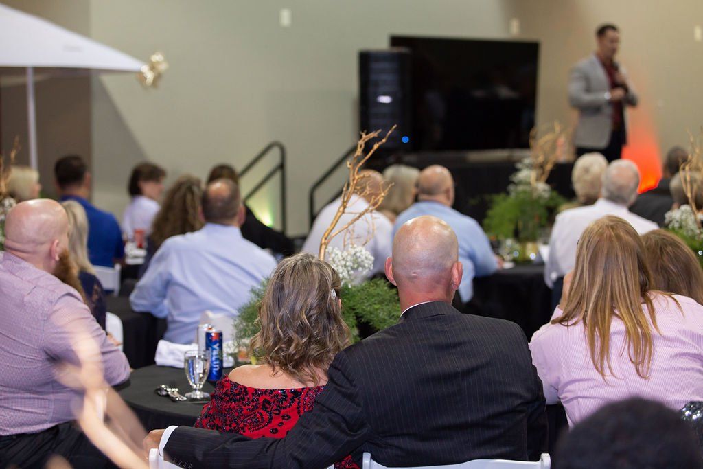 A speaker addresses an audience at a seated event, viewed from behind guests at round tables.