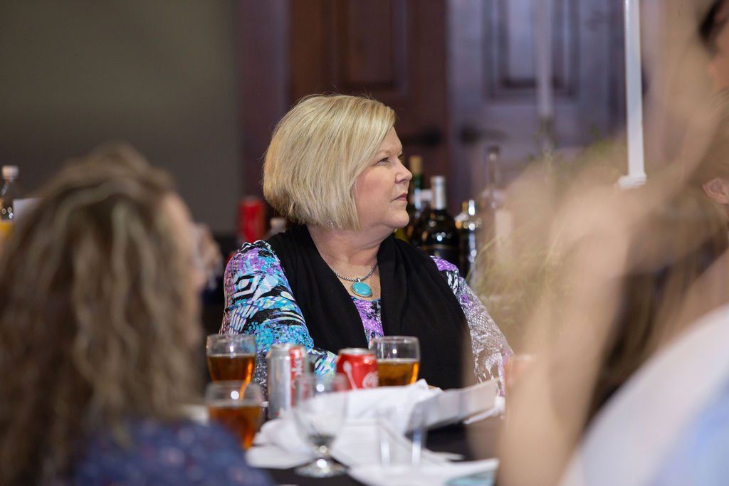 A person with short blond hair sits at a table with drinks and napkins during an indoor gathering.