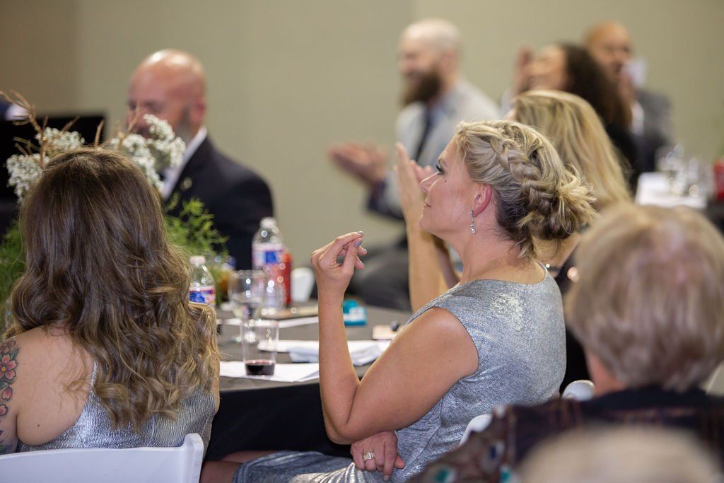 People seated at round tables in a banquet hall, listening and gesturing during an event.