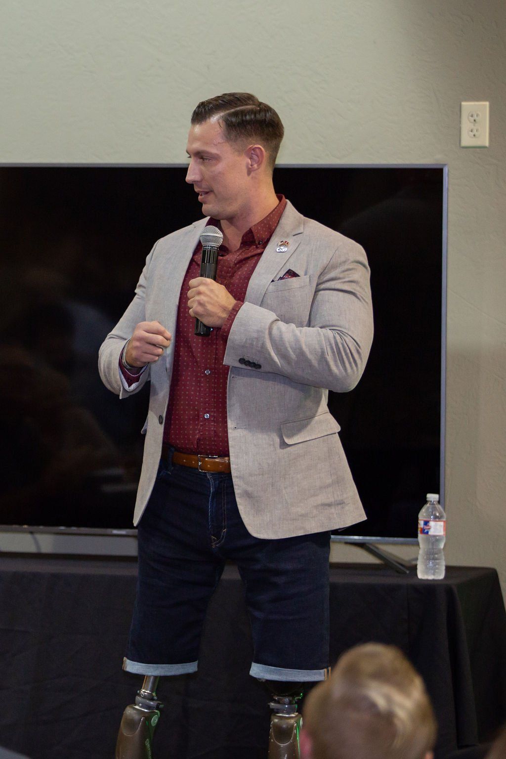 A person with prosthetic legs stands in front of a television, speaking into a microphone while wearing a blazer and shorts.