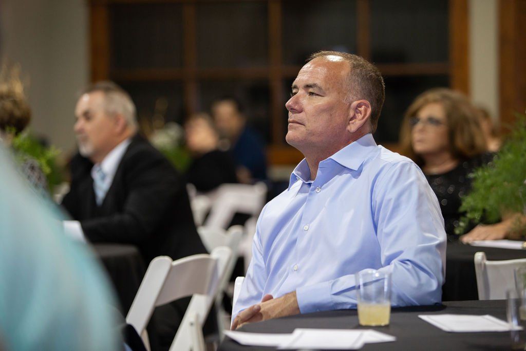 A person in a light blue button-down shirt sits at a table during an indoor event, looking toward the speaker.