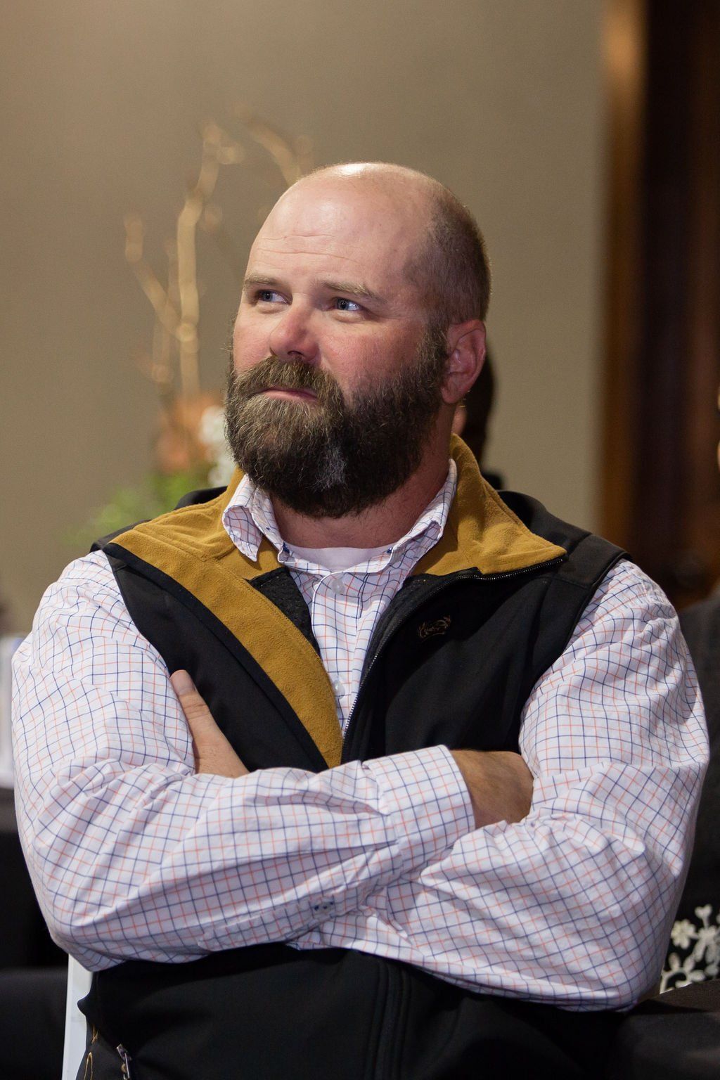 A person with a beard and bald head wears a plaid shirt and dark vest with arms crossed, looking toward the side.