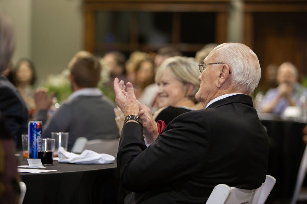 People sitting at tables at an indoor event, with a focus on an individual in a dark suit applauding.