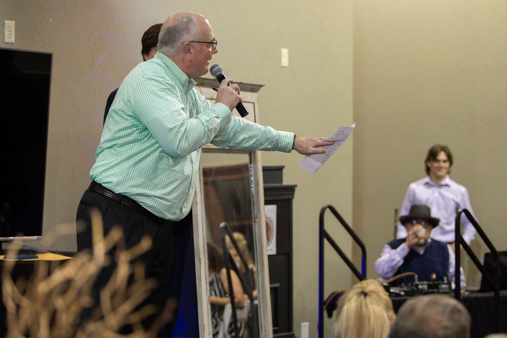 A man in a green checkered shirt speaks into a microphone while holding a paper, with others present in a room.