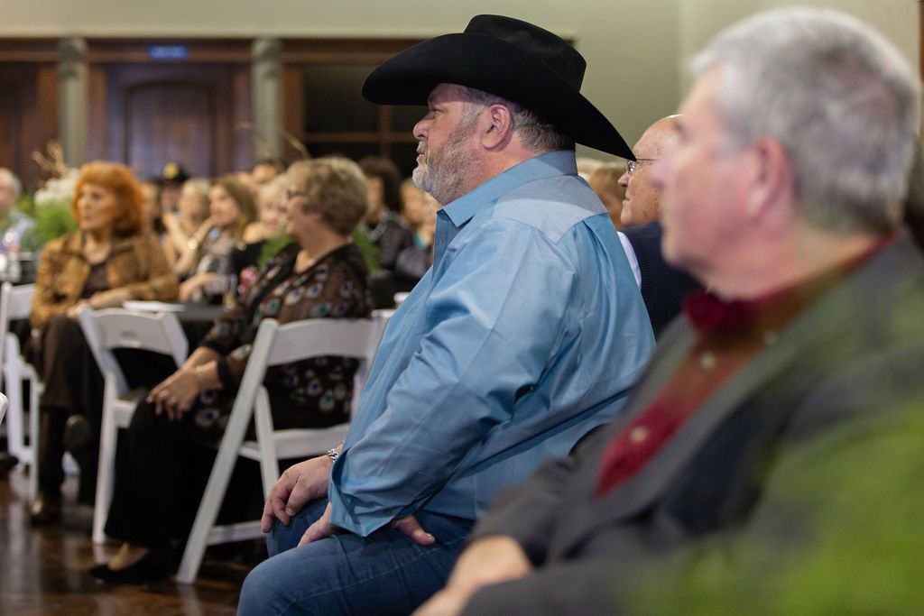 People sitting in chairs at an indoor event, focused on an unseen presentation, with a man in a cowboy hat in the foreground.