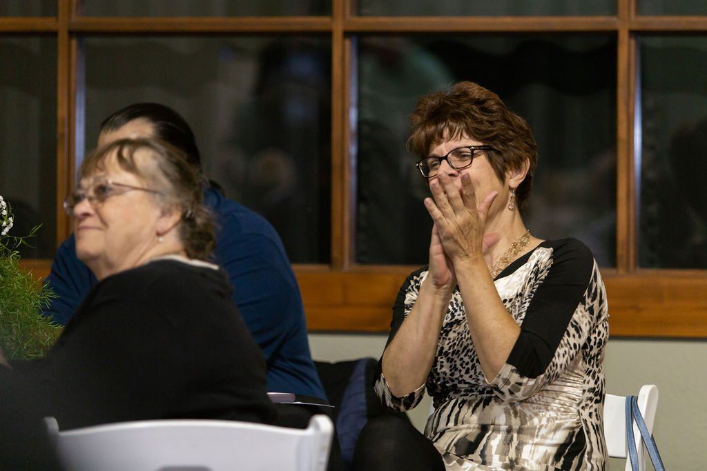 Two individuals are sitting at tables in an indoor setting; one person on the right has their hands clasped to their face.