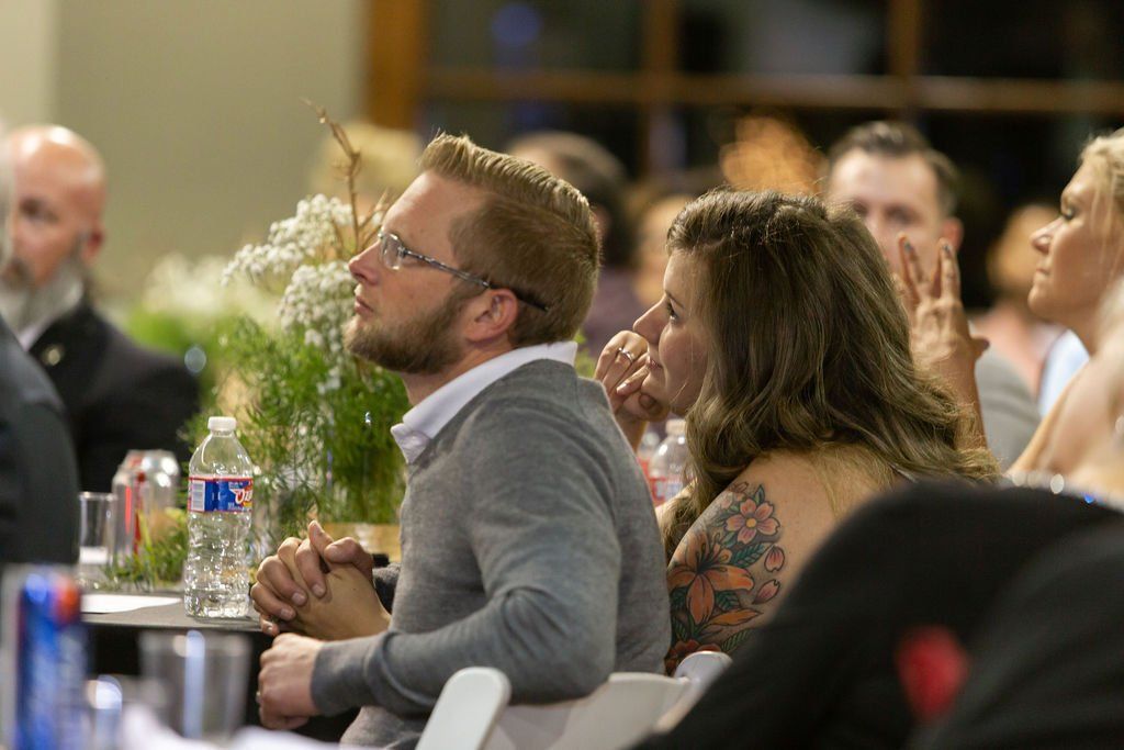 People seated at tables in a dimly lit room, watching an event with attentive expressions.