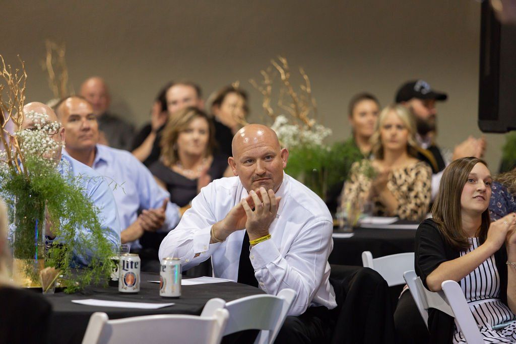 Attendees at a banquet sit at tables, some clapping, while a person in a white shirt sits centrally with chin on hands.