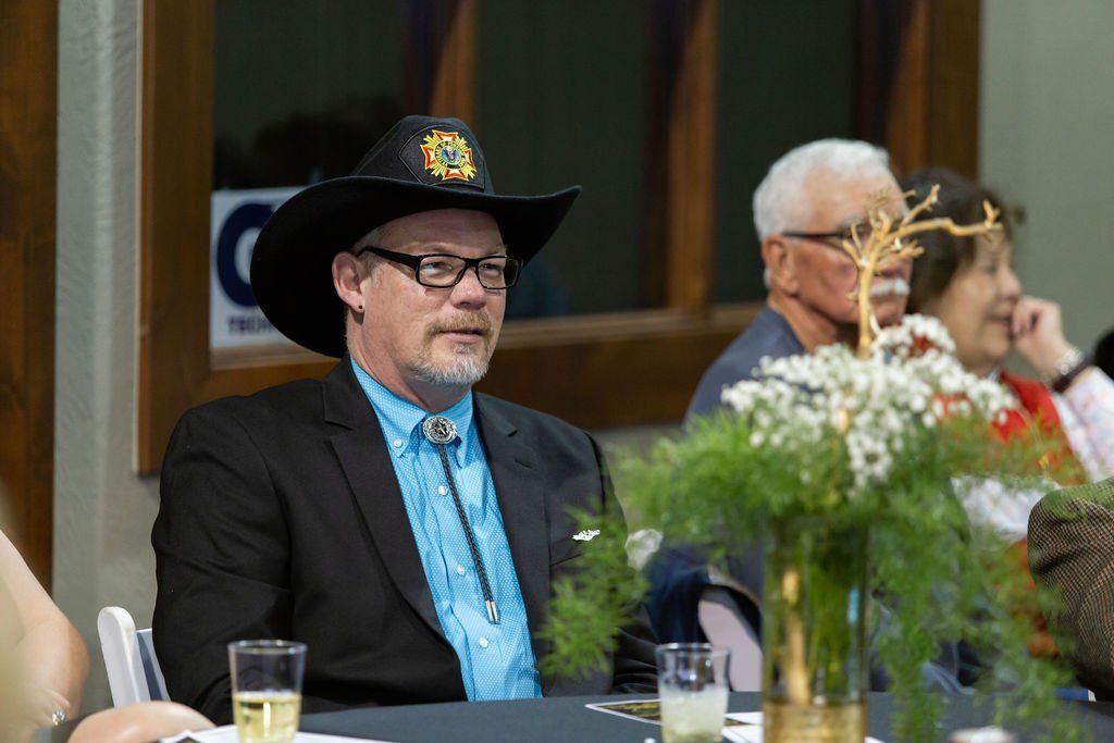 A man in a suit, bolo tie, and cowboy hat sits at a table near a floral centerpiece, with two others behind him.