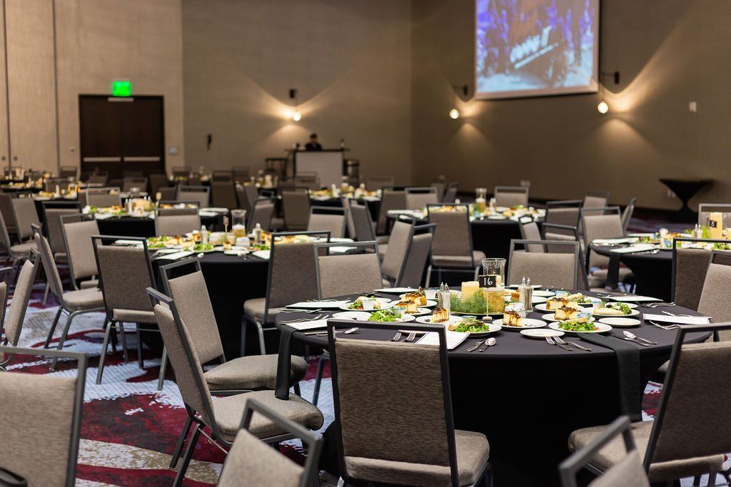 A conference room set with multiple round dining tables draped in black cloths and gray chairs on patterned carpeting.