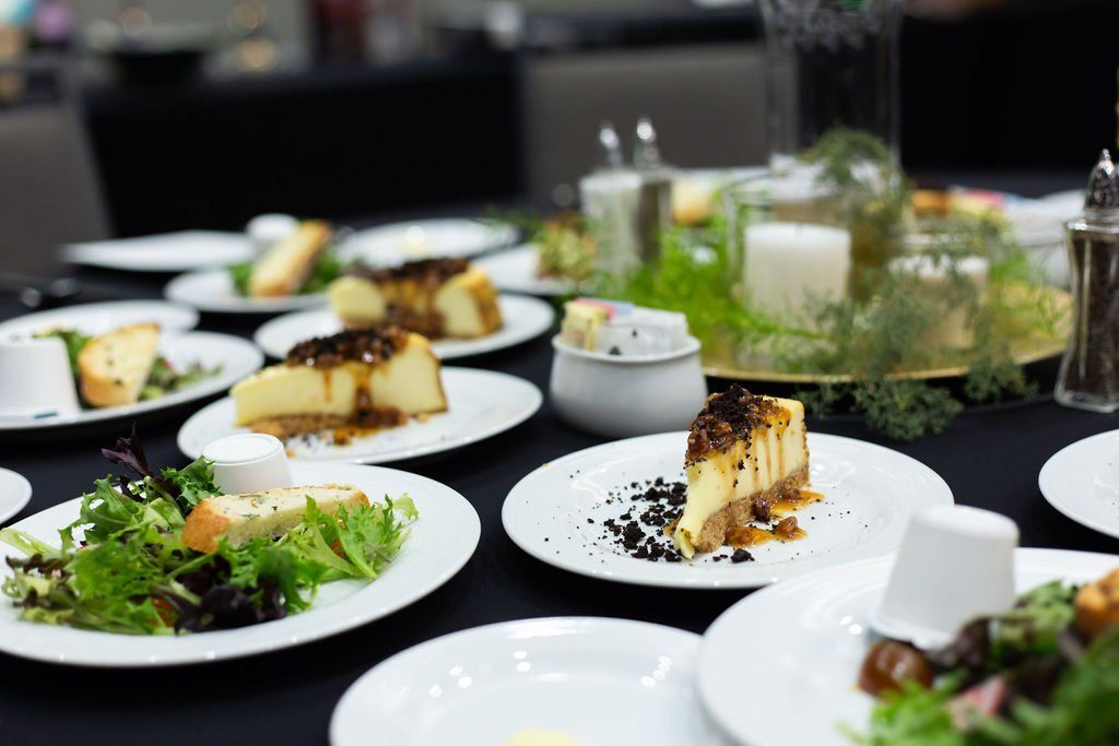 Multiple plates of cheesecake and salad are arranged on a dark tablecloth, with a floral centerpiece in the background.