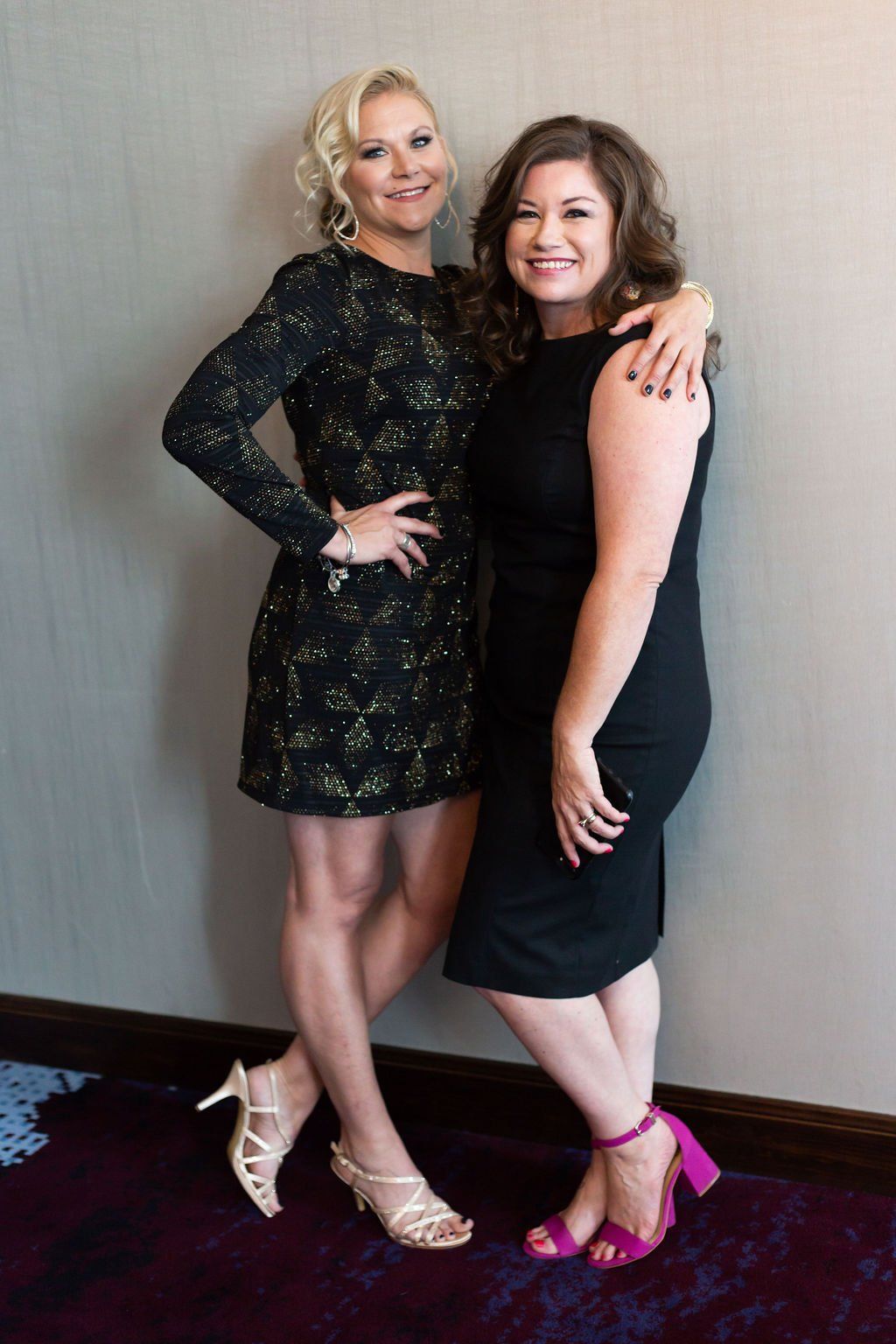 Two women in black dresses pose together smiling, one with a hand on the other's shoulder, standing against a wall.