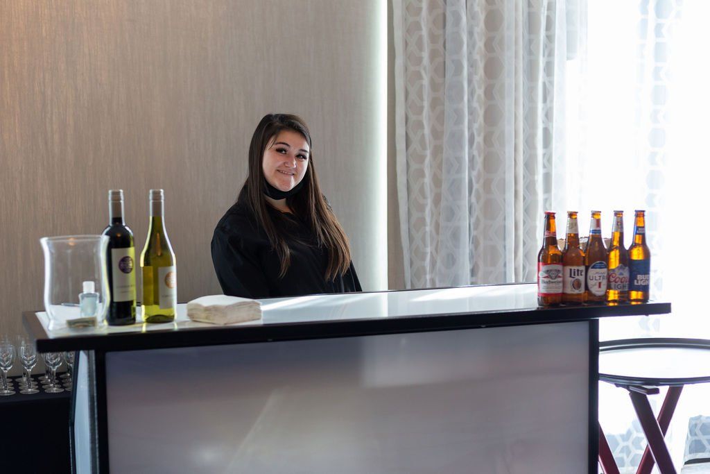 A person stands behind a lit, white bar stocked with wine bottles and beers in an indoor room with light-colored curtains.