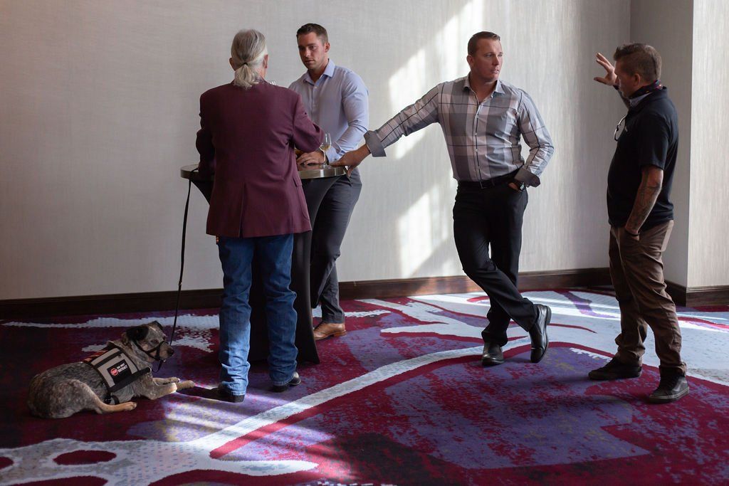 Four people stand in a room with a patterned rug; one person gestures while another has a service dog resting nearby.