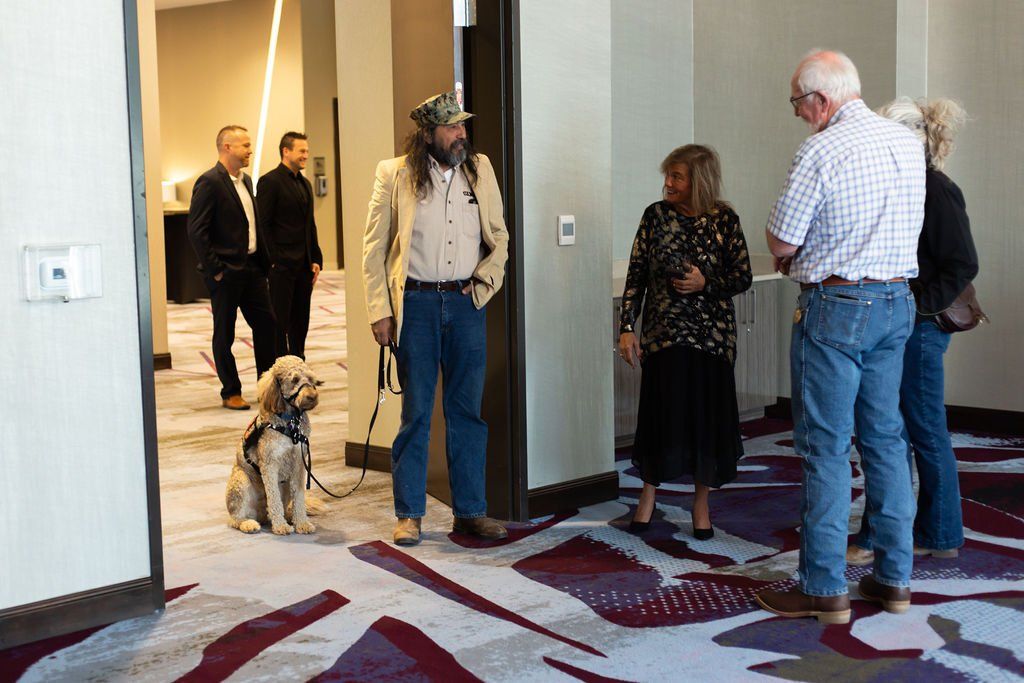 A group of people standing and talking in a hallway, with a dog sitting near a man in a beige jacket.