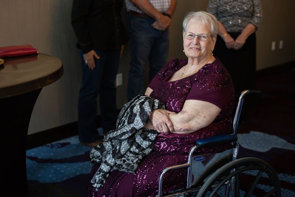 A person in a sparkling dark purple dress sits in a wheelchair at an indoor event, with others standing behind them.