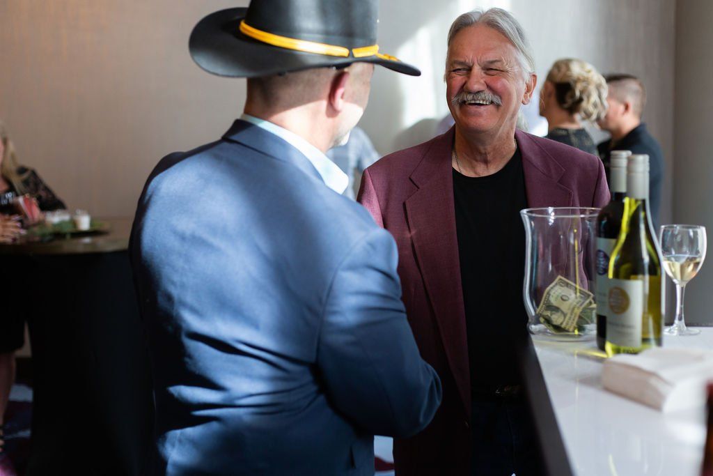 A person in a cowboy hat and blue suit shakes hands with a smiling person in a maroon jacket at a bar.