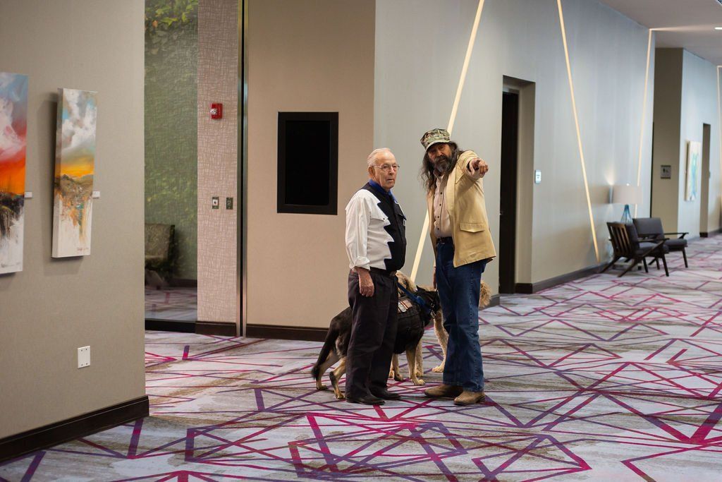 Two people stand in a hotel hallway with a dog, one pointing towards an open doorway.