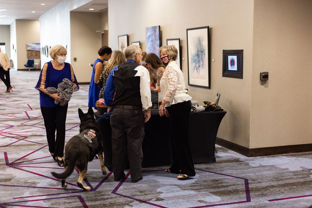 People gather in a hallway to view art displayed on a wall, while a person walks a German Shepherd toward them.