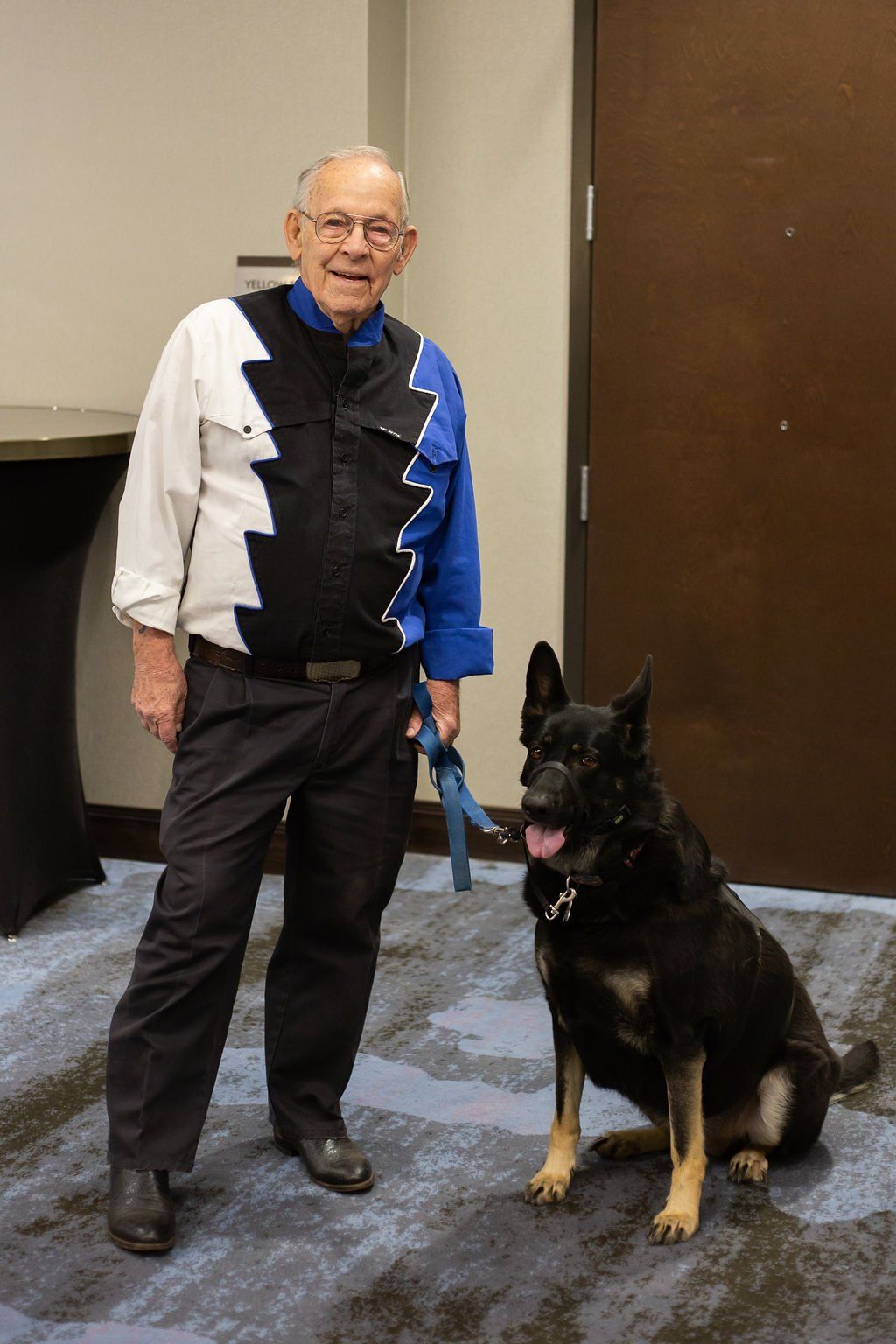 A person with glasses stands beside a large black and tan German Shepherd sitting on a blue patterned carpet indoors.
