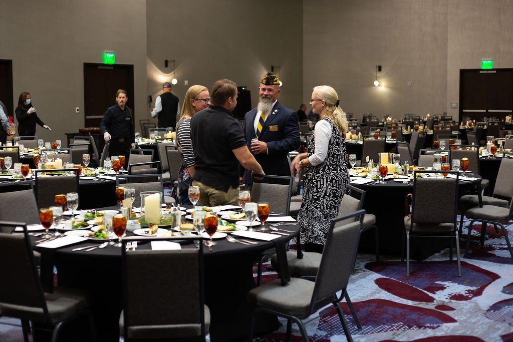 Attendees in formal attire converse around banquet tables in a spacious, dimly lit conference hall.