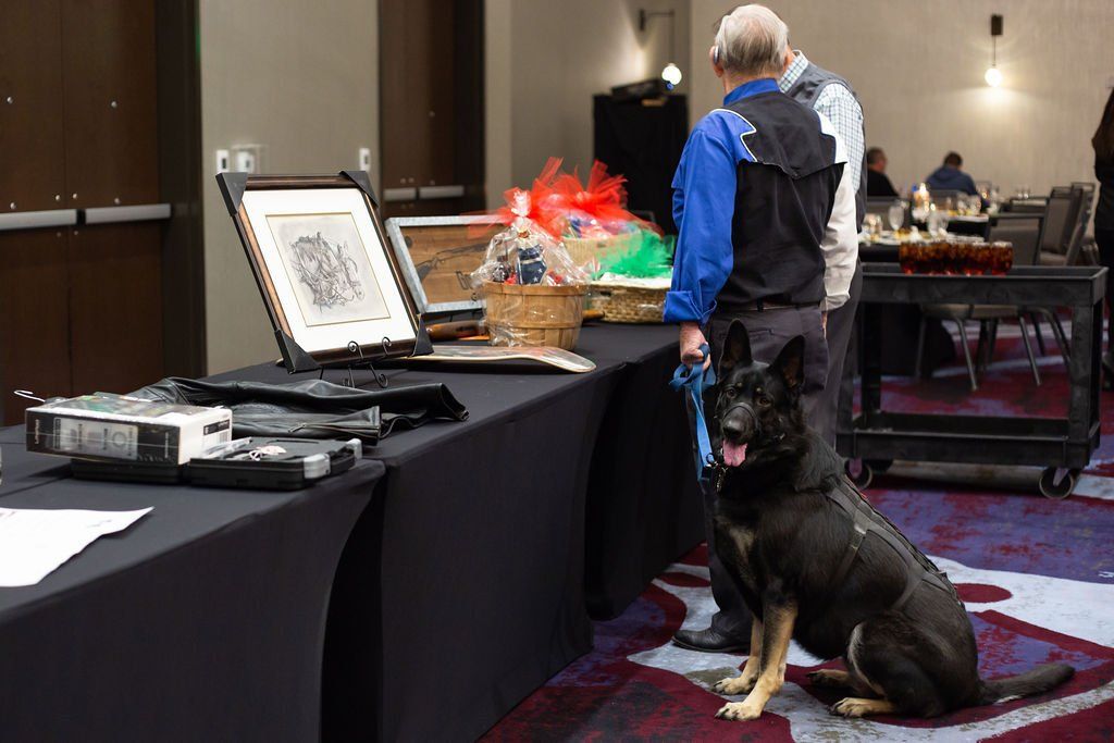 A German Shepherd service dog sits by its handler at a table featuring auction items in a hotel event space.