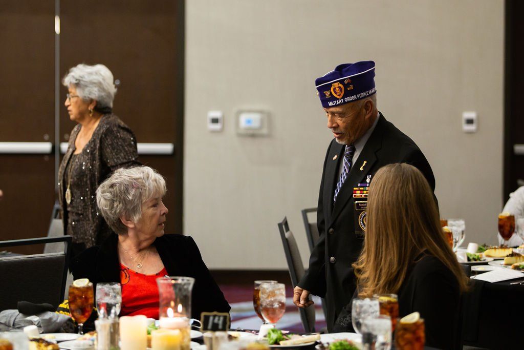 An individual in a military-style uniform and cap walks past a seated person at a banquet table.