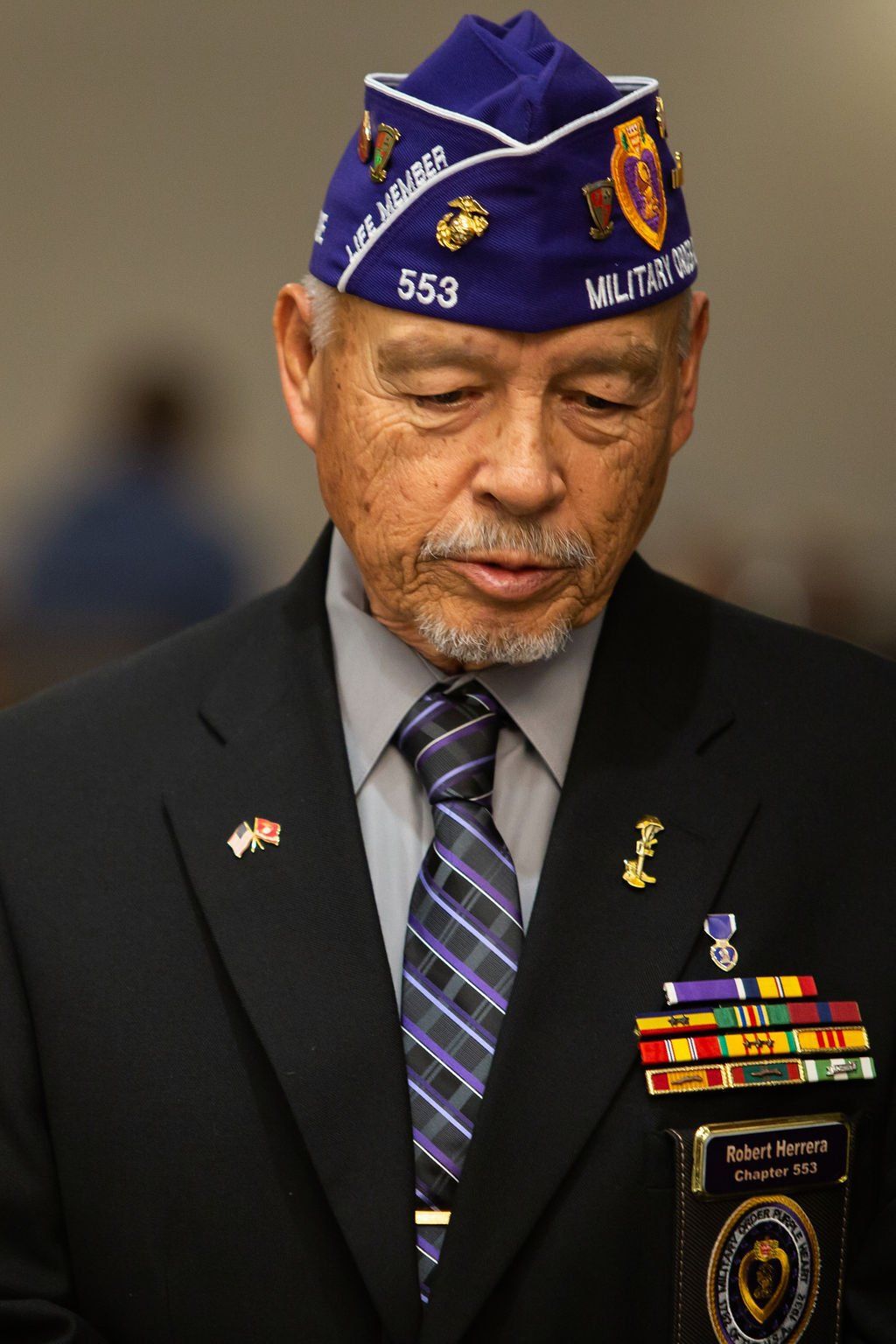 A veteran wearing a military dress cap and suit with medals and ribbons pinned to his jacket, head bowed in reflection.