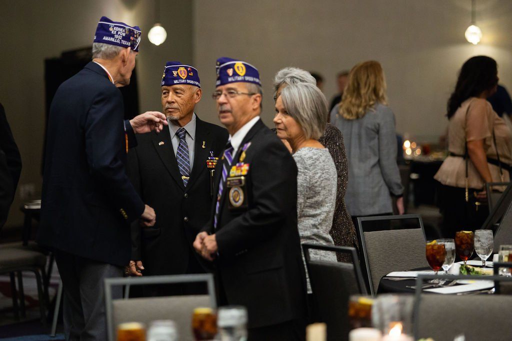 Four people in formal attire and veteran caps socialize in a banquet hall, with tables and guests visible in the background.