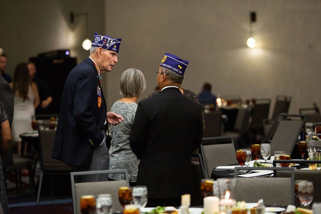Two people in formal attire and veteran caps stand talking in a banquet hall with tables and chairs in the background.