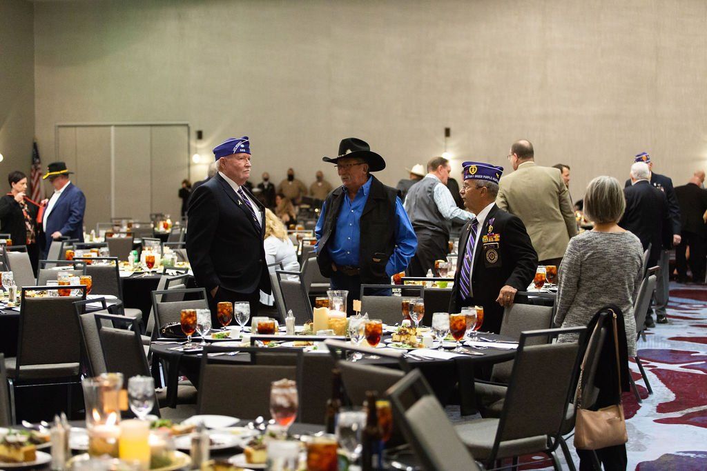 People in casual and formal attire, some wearing veteran caps, gather at tables in a large hotel event room.