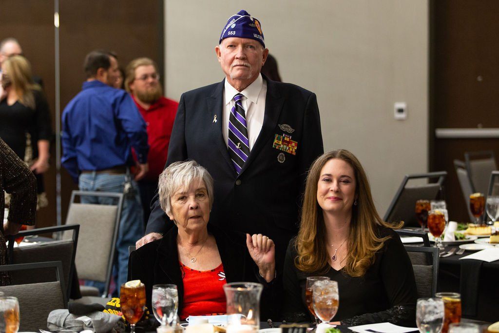 A man in a suit and service cap stands between two seated individuals at a banquet, with guests in the background.