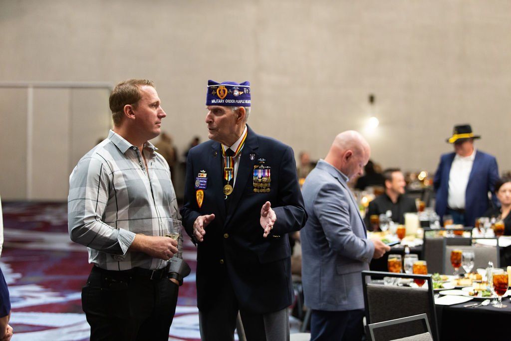 Two men converse in a conference room; one wears a veteran’s cap and blazer, the other a patterned dress shirt.
