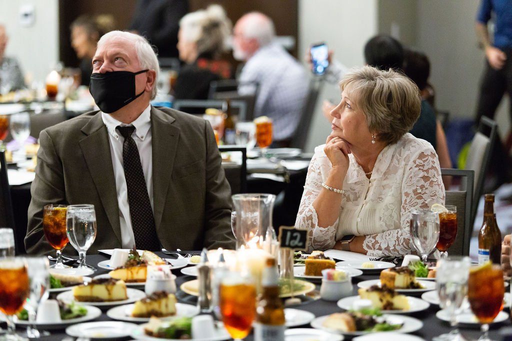 A man in a suit and mask and a woman in a white lace top sit at a dinner table looking upward, surrounded by served plates.