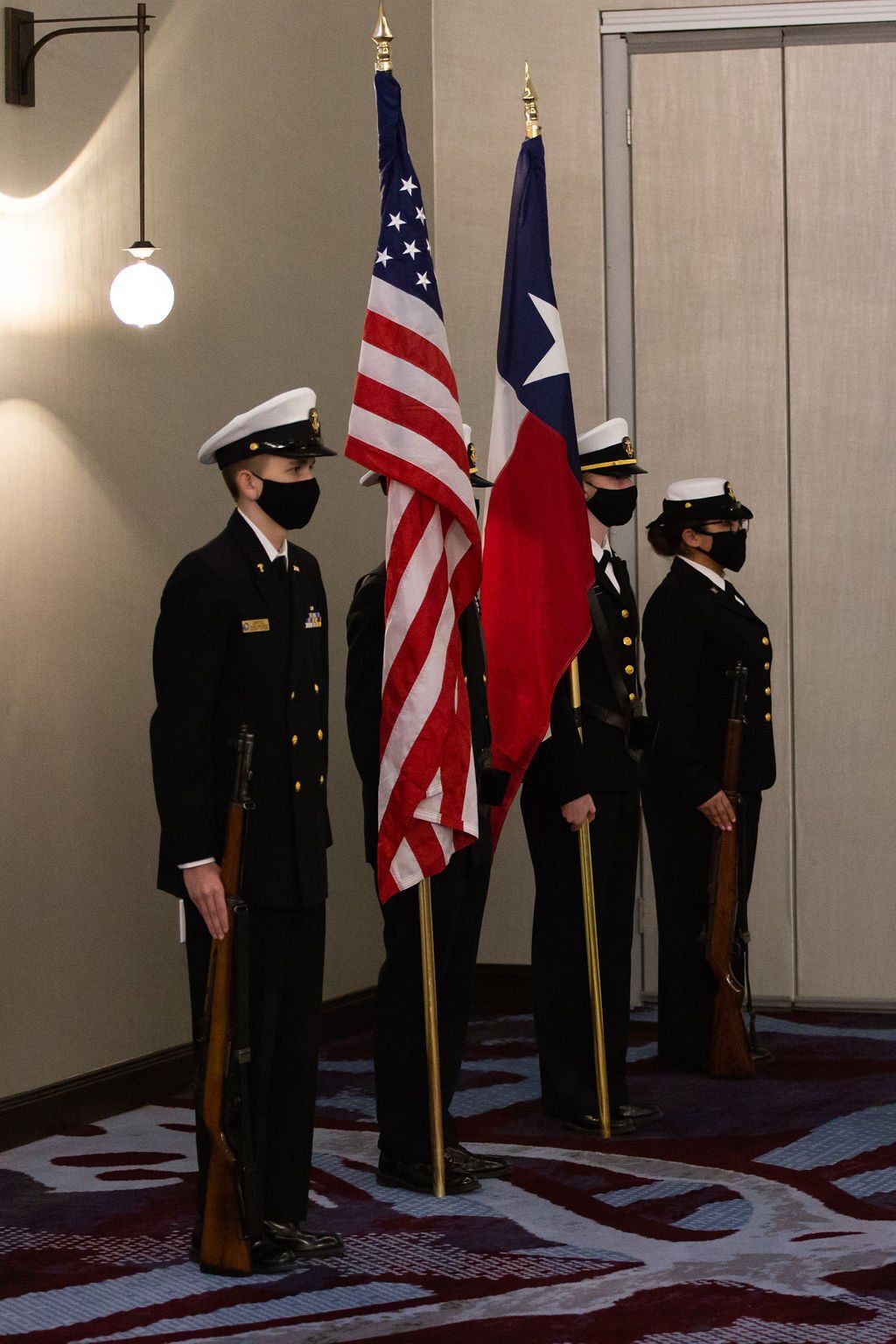 Four individuals in military-style dress uniforms and face masks stand in a line holding ceremonial rifles and two flags.