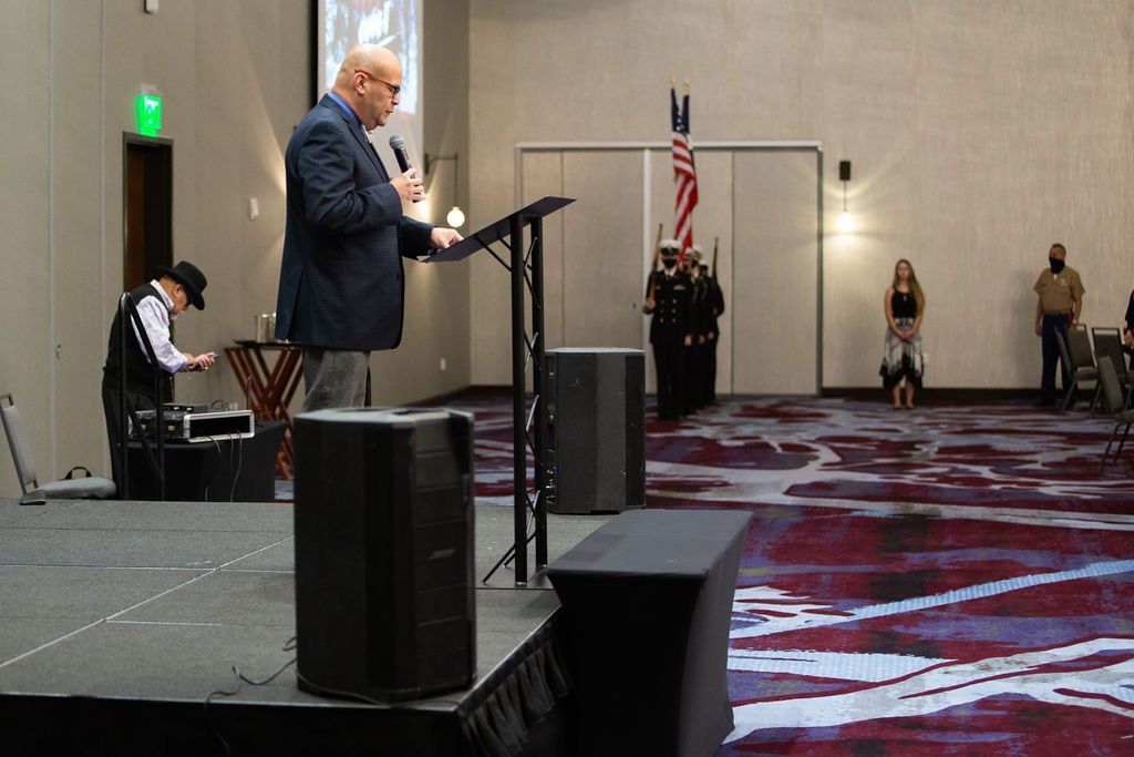 A speaker at a podium in a hotel ballroom, with a person operating audio equipment and a color guard in the background.