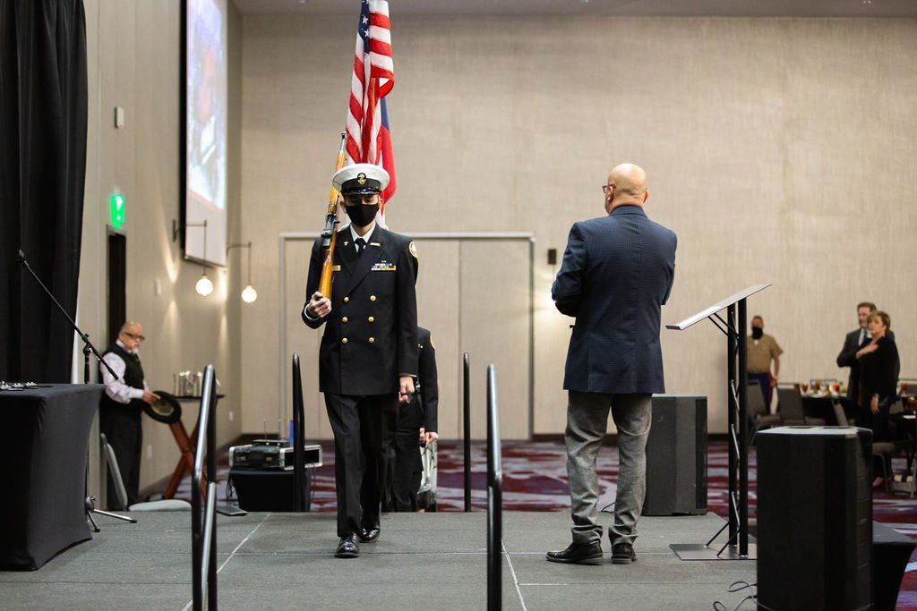 A uniformed person carries a flag across a stage while a person in a suit watches, inside a large meeting hall.