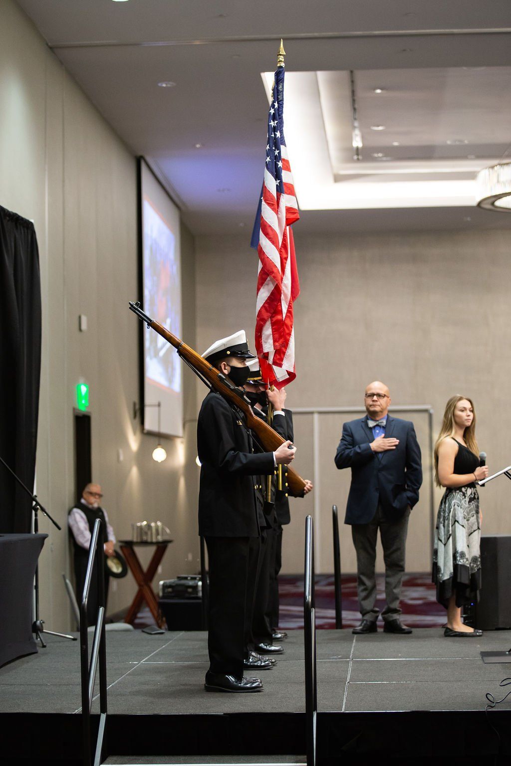 A color guard holds an American flag and rifle on stage during an event, while two individuals stand with hands over hearts.